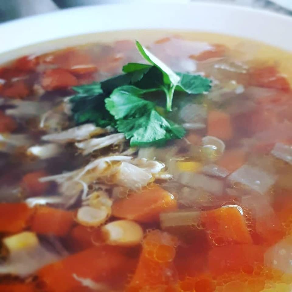 Clear chicken soup with carrots and corn in a shallow white bowl. Garnished with flat leaved parsley.