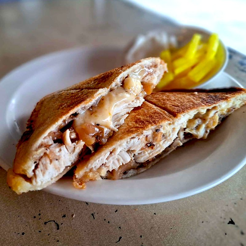 Hot jaffle cut in half with one half on top of the other. Filing is exposed. Chicken breast, melted cheese and fruit chutney. Accompaniments are bright yellow batons of pickled daikon in turmeric.
