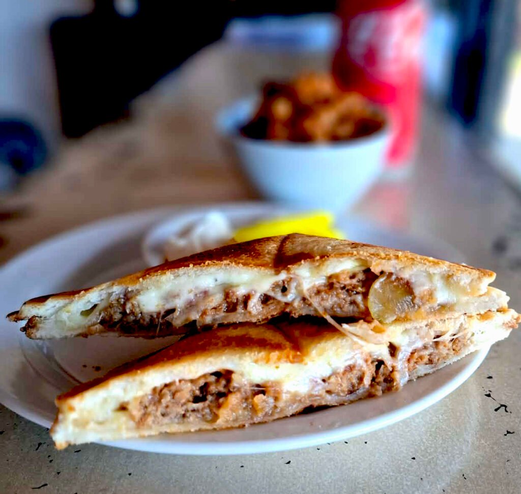 Cheeseburger jaffle cut in half diagonally, halves stacked showing melted cheese and beef filling. Bowl of baked beans and can of Coke in the background.