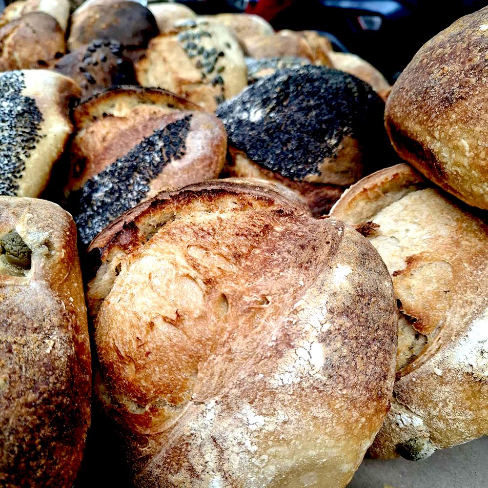A pile of sourdough loaves in various bulbous shapes, some with seeds on top and some with flour dusted on top. These sourdough loaves are crusty and brown bulging toward the viewer.